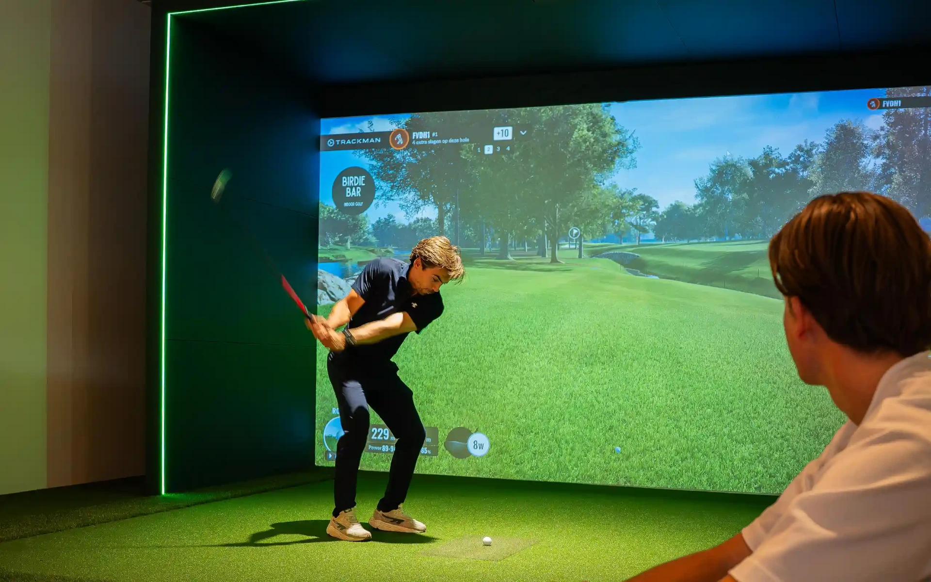 Golfer slaat af op de golfsimulator terwijl een vriend toekijkt